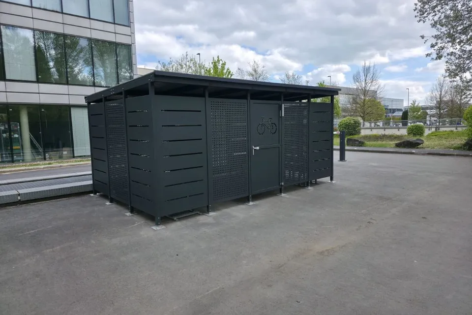 Abri vélos fermé Optimo en acier gris anthracite, parois ajourées et porte sécurisée à pictogramme vélo, installé sur le parvis d'un immeuble de bureaux.